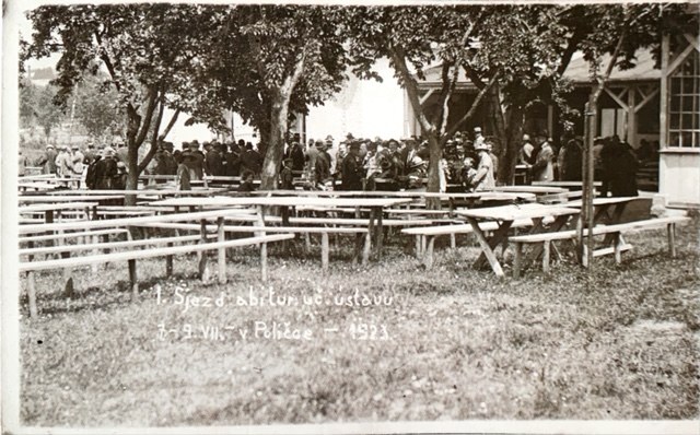 Dav slavnostně oblečených lidí mezi stoly a lavicemi v zahradě lázní, fotografie z roku 1923