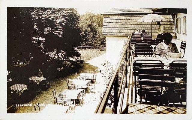 Fotografie terasy letoviska: řady stolů s kostkovanými ubrusy, slunečník a hosté, pod terasou zahrada