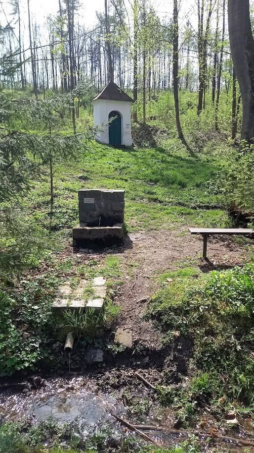 Kamenná studánka pod kapličkou, vedle lavička a dřevěná lávka