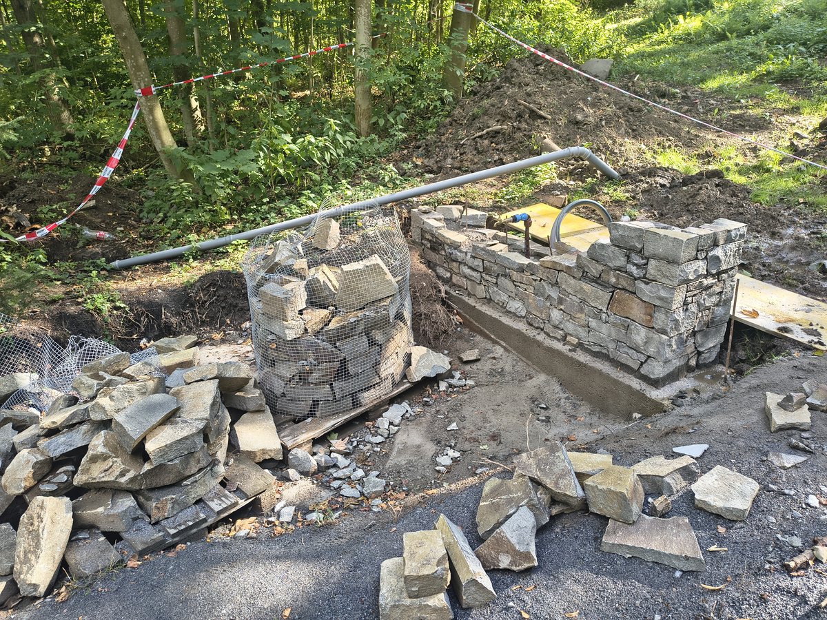Rozestavěná kamenná zídka studánky, kolem palety s kamenem a vytyčovací páska