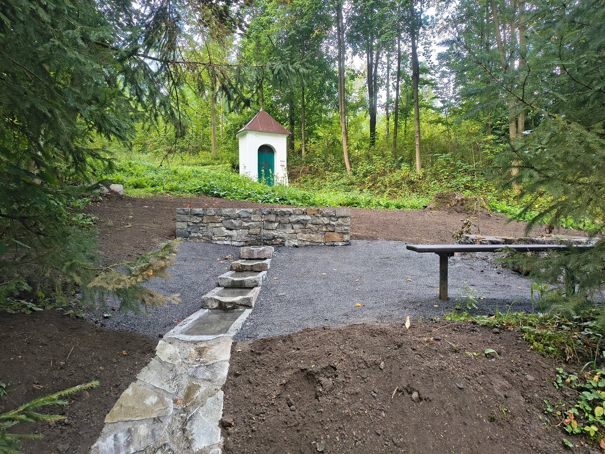 Nové okolí studánky: kamenná zídka, koryto s kaskádou, mlatová plocha a lavička, v pozadí kaplička